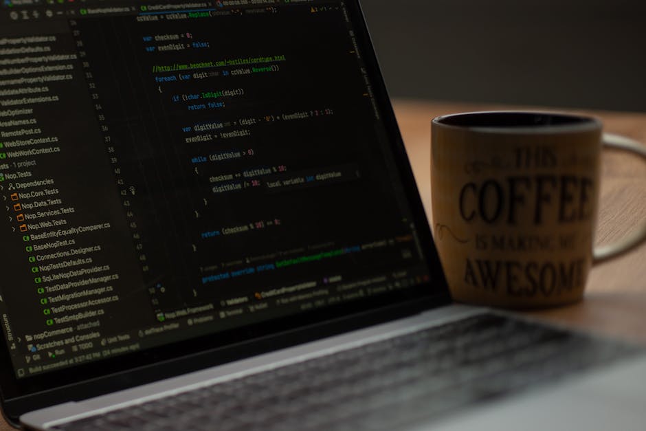 A close-up photograph showing a laptop on a wooden surface with visible lines of colorful programming code displayed on its screen. Next to the laptop, there is a ceramic coffee mug with a beige backg