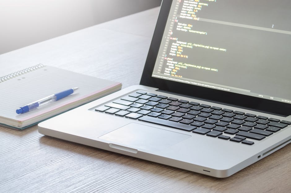 A close-up view of a silver laptop placed on a wooden desk displaying lines of code on its screen. Next to the laptop, there is a spiral-bound notepad with a blue pen resting on top, indicating a work